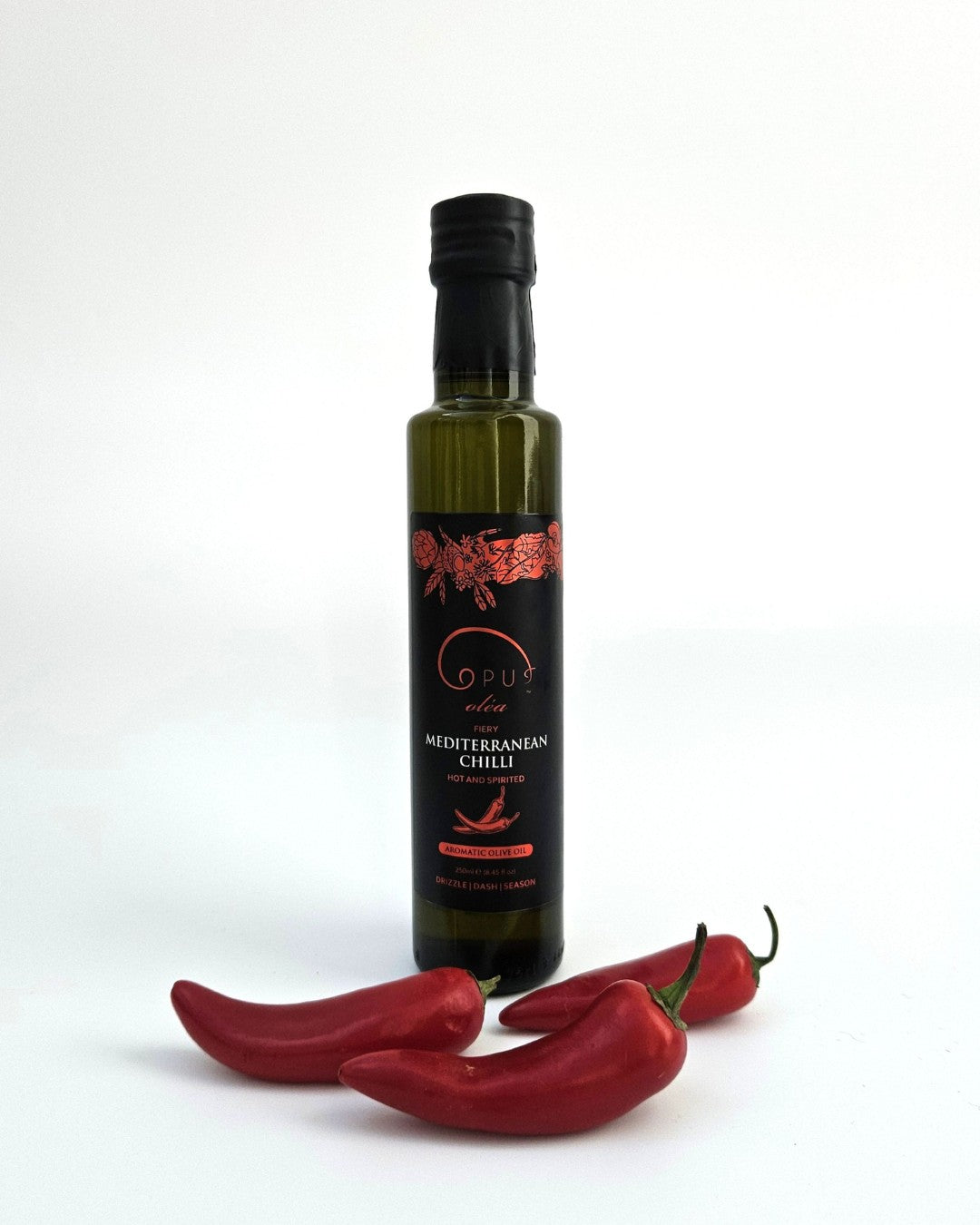 A bottle of Opus Mediterranean Chilli olive oil styled with three whole red chilli peppers at its base.