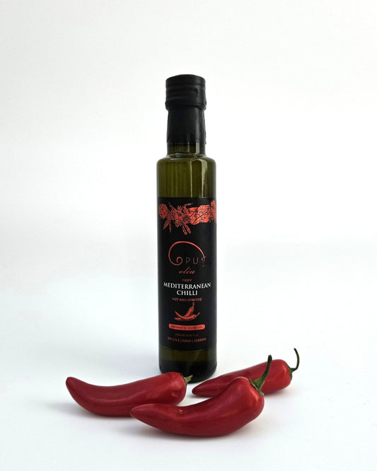 A bottle of Opus Mediterranean Chilli olive oil styled with three whole red chilli peppers at its base.